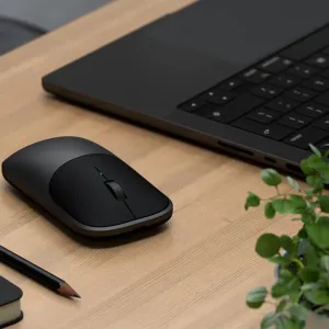 Satechi Slim EX trådløs mus på et skrivebord ved siden av en bærbar PC, med lavprofilert svart design og rullehjul.