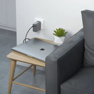 Cargador de pared Satechi de 100 W GaN PD enchufado junto a un portátil, con un cable USB-C trenzado sobre una mesa auxiliar.