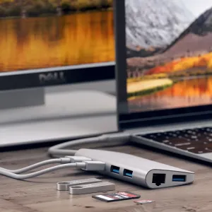Hopeanvärinen Satechi USB-C-multimediasovitin, jossa on USB-portteja ja Ethernet-liitäntä, sekä SD-kortteja ja kaapeleita pöydällä