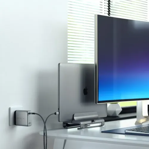 Satechi vegglader i sølv med USB-C-port, plugget inn i stikkontakten og koblet med flettet kabel til den bærbare datamaskinen på skrivebordet.