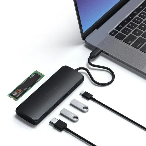 Satechi USB-C-hub i svart ligger koblet til den bærbare datamaskinen; siden med flere USB-A-porter og HDMI, omgitt av kabler og minner.