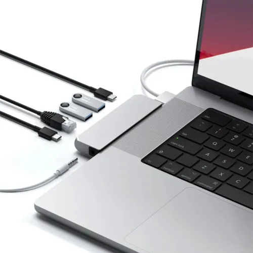 Satechi Pro Hub Mini in silver connected to MacBook, with visible headphone jack; next to it are cables and USB sticks on table