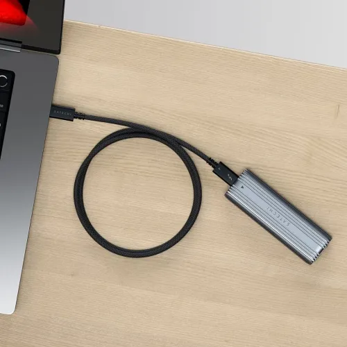 Musta punottu Thunderbolt-kaapeli USB-C:llä, joka on liitetty kannettavaan tietokoneeseen, pyöreä silmukka ja harmaa pitkulainen kotelo puupöydällä.