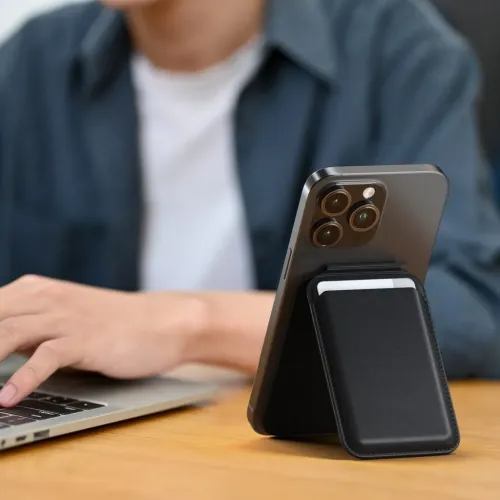 Juodas magnetinis piniginės stovas, pritvirtintas prie "iPhone" nugarėlės, sulankstytas kaip atrama su kišenėje matoma kortele, stovi ant stalo.
