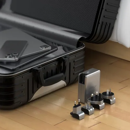 Satechi USB-C GaN-reiselader i sølv står på tregulv med fire utskiftbare reiseadaptere foran en åpen koffert, ved sengen.