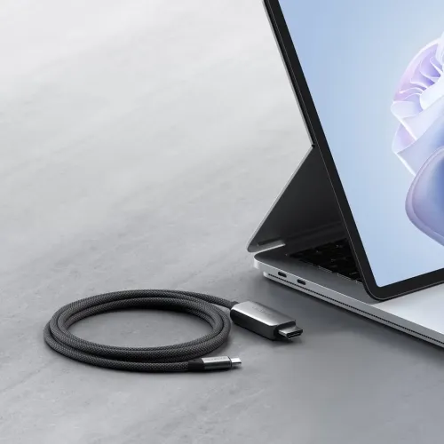 Satechi USB-C-HDMI kabelis ar USB-C un HDMI savienotājiem melnā pītā dizainā, sarullēts blakus klēpjdatoram uz galda virsmas.