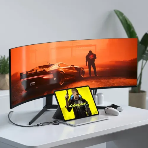 USB-C til HDMI-adapter med flettet kabel og HDMI-port, i mørkt metallhus, på skrivebordet mellom nettbrettet og den buede skjermen.