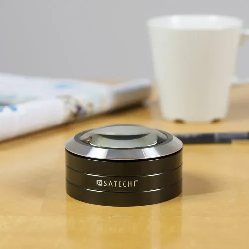 Satechi Read Mate forstørrelsesglass i rund, svart aluminium med kuppelformet linse på bordet, logo på forsiden; uskarpt krus og magasin.