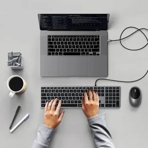 Satechi W3 USB-C-tastatur med numerisk tastatur, vist tilsluttet med kabel til en bærbar computer på et skrivebord, mens hænder skriver