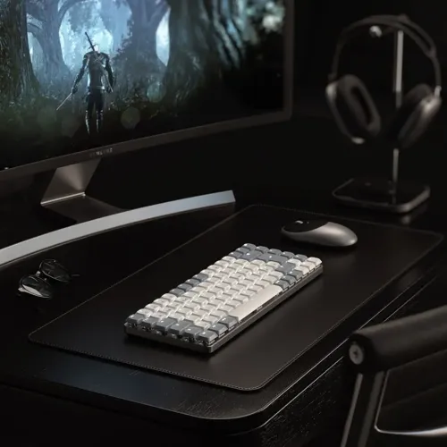 Ljusgrått kompakt tangentbord med tvåfärgade tangenter på svart skrivbordsmatta, placerat bredvid trådlös mus vid skärm.