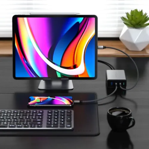 Satechi 100 W GaN ceļojumu lādētājs ar USB-C un USB-A pieslēgvietām uz galda darbina planšetdatoru un viedtālruni.