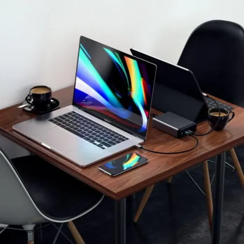 Reiselader i mørkegrått med porter på kortsiden av skrivebordet, koblet med kabel til bærbar PC og nettbrett, ved siden av mobilen.