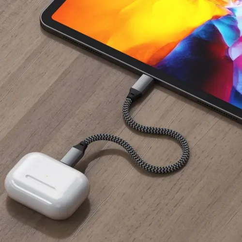Lyhyt punottu USB-C-Lightning-kaapeli, jonka väri on avaruudenharmaa, yhdistää tabletin valkoiseen kuulokekoteloon vaaleanruskealla puupöydällä.