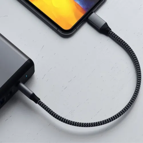 Satechin lyhyt harmaa punottu kaapeli, joka on liitetty tumman virtalähteen USB-C-portin ja vaalealla puupinnalla olevan älypuhelimen välille.
