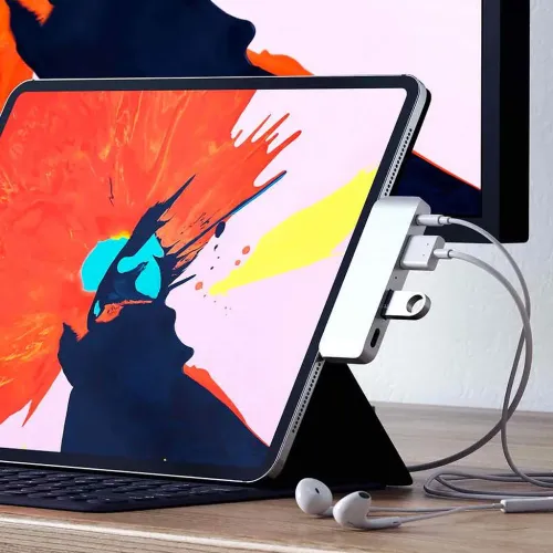 Sølvfarvet USB-C-hub monteret på iPad Pro med tilsluttede kabler, synlige USB- og 3,5 mm-porte og ekstern skærm i baggrunden.