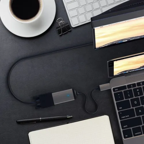 Satechi USB-C til VGA-adapter i space grey med blå LED og kort kabel, tilsluttet mellem bærbar USB-C og VGA-kabel på bordet.