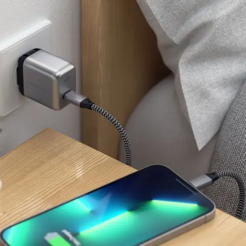 Satechi USB-C väggladdare med USB-C-port, ansluten i vägguttag, laddar en telefon via flätad kabel på nattduksbord av trä.