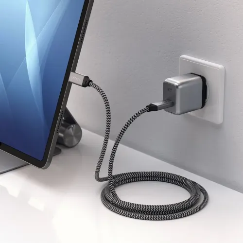 Satechi USB‑C väggladdare i aluminium sitter i uttag, med flätad USB‑C‑kabel ansluten till surfplatta på vitt skrivbord.