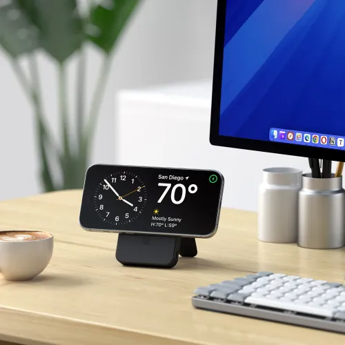 Svart powerbank med integrert stativ som holder smarttelefonen horisontalt på skrivebordet, foran skjerm, tastatur og kopp.