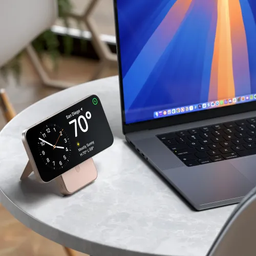 Magnetisk powerbank med stativ i ørkenrosa holder en smarttelefon horisontalt; USB-C-porten er synlig; på bordet ved siden av en grå bærbar PC.