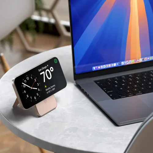 Powerbank med innebygd stativ i Desert Rose holder en smarttelefon horisontalt på et rundt bord ved siden av en bærbar PC.