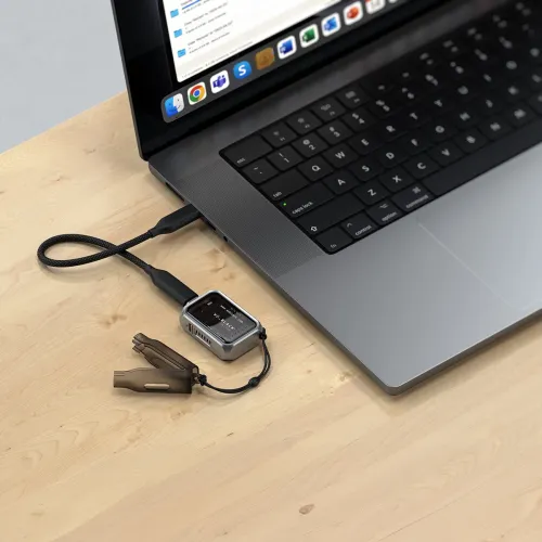 Mini NVMe SSD-kabinett i metall med transparent fönster, anslutet med kort USB‑C‑kabel till laptop på träbord, med nyckelrem