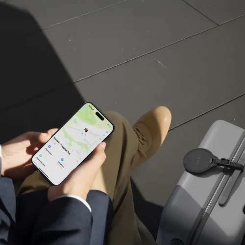 Svart, rund bagasjelapp i vegansk lær med spenne, festet på den grå kofferten nær glidelåsen; Satechi-logoen er synlig på toppen.