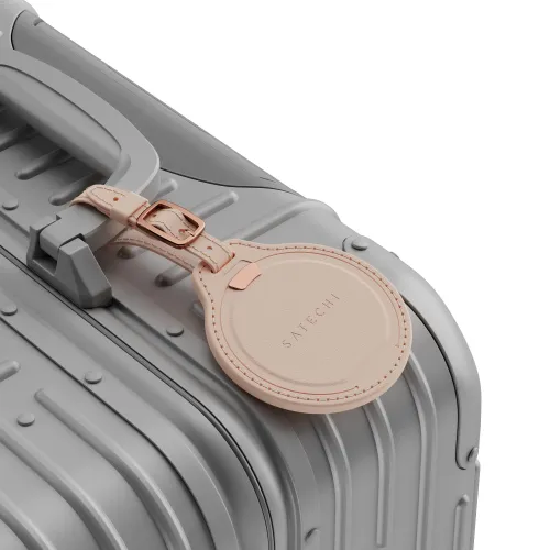 Rund bagasjelapp i lys rosa vegansk lær fra Satechi med spennestropp, festet til håndtaket på en grå hard koffert med ribbestrikk.