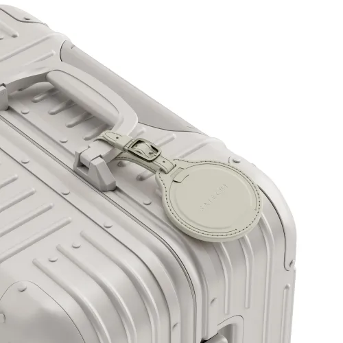 Rund, sandfarget bagasjelapp i skinnlook fra Satechi, med preget logo og stropp med spenne, festet til håndtaket på kofferten.