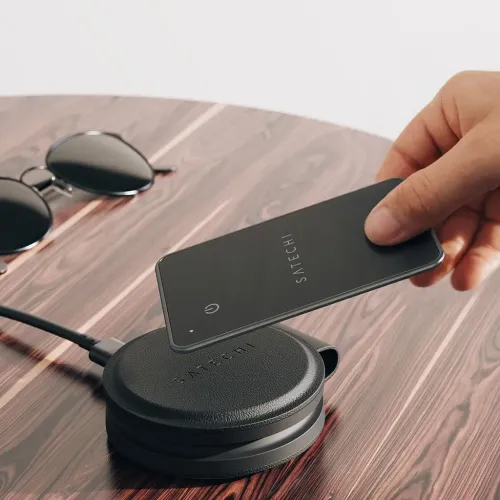 "Satechi FindAll" piniginė kortelė, laikoma virš apvalios įkrovimo plokštelės ant medinio stalo; itin plona kortelė su "Satechi" logotipu ir mygtuku.