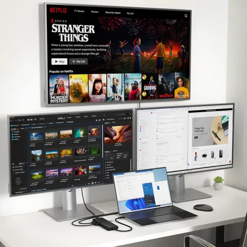 Melns USB-C daudzvadu adapteris uz balta galda, taisnstūra formas, ar vairākiem kabeļiem, kas savienoti ar klēpjdatoru un diviem ārējiem monitoriem.