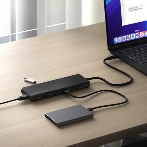 Satechi USB-C daudzportu adapteris melnā krāsā uz darbvirsmas, pievienots klēpjdatoram, ar pieslēgtām pieslēgvietām, kabeļiem un ārējo SSD disku.