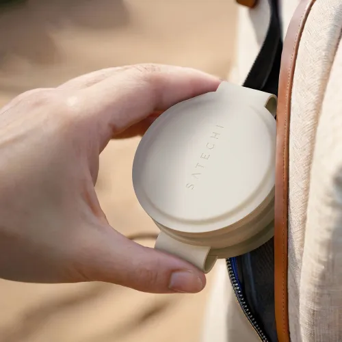 Sandfarget, rund Satechi-lader med SATECHI-logo, som holdes mellom fingrene og legges i en lomme med glidelås i en veske.