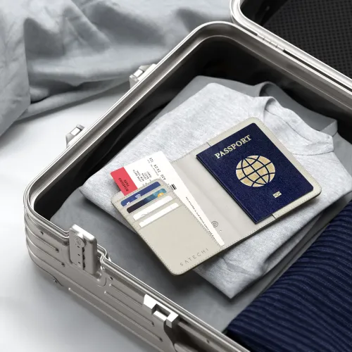 Sandfarget Satechi-passetui ligger åpent i en sølvfarget koffert, med kortspor, boardingkort og pass på grå skjorte.