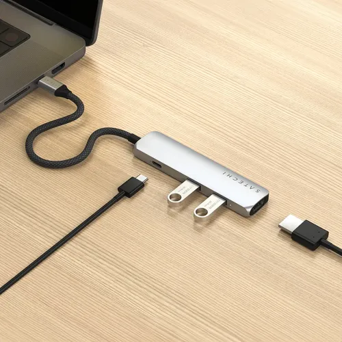 Hopeinen Satechi USB-C -moniporttisovitin liitettynä kannettavaan tietokoneeseen, punottu kaapeli puupöydällä.